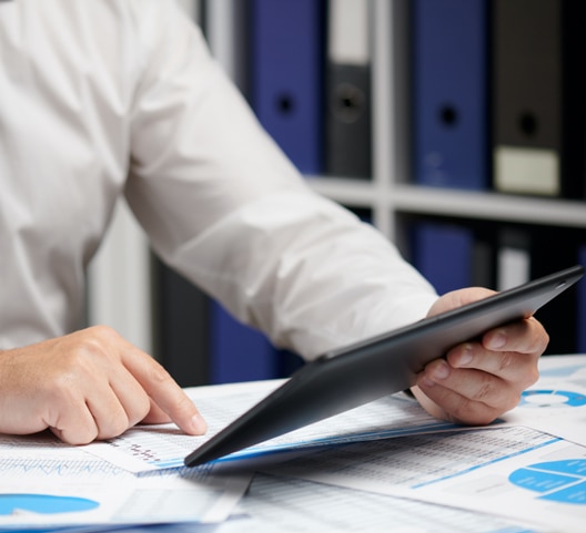 A person in a white shirt is using a tablet at a desk, surrounded by documents and binders. Their focus is on the tablet screen, with one hand pointing at printed data. The background features shelves filled with blue and black binders.