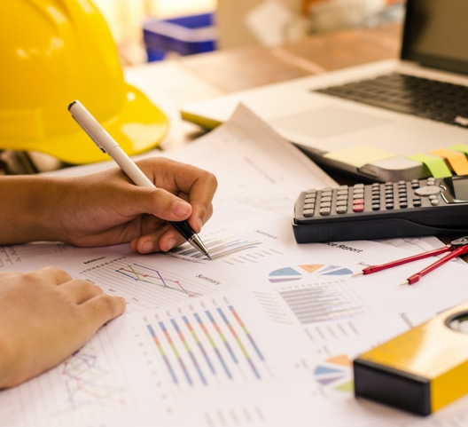 A person writing on graphs and charts spread on a table, with a calculator, red measuring tape, and a yellow hard hat nearby. A laptop is partially visible, and the scene suggests a work environment involving data analysis or engineering.