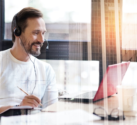 A man wearing a headset sits at a desk, smiling and looking at a laptop. He holds a pen and writes on paper, suggesting he is engaged in an online meeting or remote work. Sunlight filters through a window in the background.