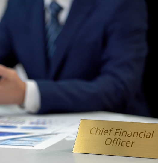 A person in a suit sitting at a desk with a gold nameplate reading "Chief Financial Officer." Documents and a blurred figure are in the background.