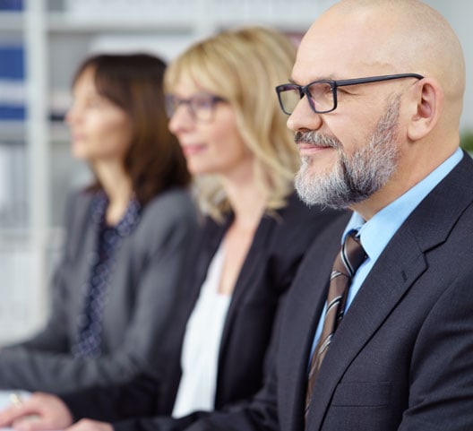 A group of three people sitting in a row, all wearing business attire. The focus is on a bald man with glasses and a beard. Two women with glasses and medium-length hair are in the background, slightly out of focus.