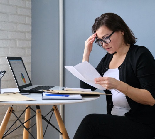 A woman sits at a round table, reading papers. She wears glasses and a black cardigan. A laptop with a colorful pie chart is open in front of her, alongside a pen and notebook. The setting appears to be a home office with a light-colored wall.