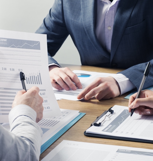 Three people in business attire sit at a table, reviewing financial reports with charts and graphs. They are holding pens and examining documents on clipboards, suggesting a business meeting or financial discussion anchored by professional bookkeeping services.