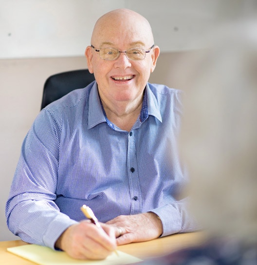 A bald man wearing glasses and a blue checkered shirt is sitting at a desk, smiling while writing about business cash flow management strategies on a yellow notepad. The background is out of focus, suggesting an office or meeting setting focused on business transformation services.