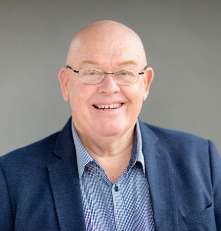 A smiling bald man wearing glasses, a blue checked shirt, and a dark blue blazer stands confidently in front of a plain gray background, ready to offer expert small business advisory services.