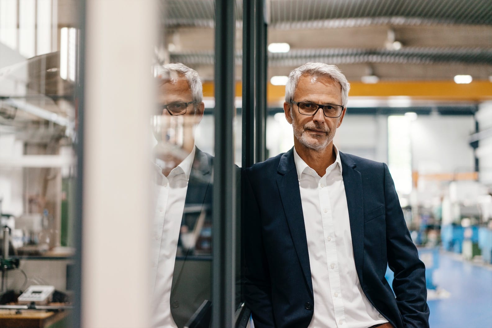 A man with gray hair and glasses is leaning against a glass wall in a modern industrial setting. He is wearing a dark suit jacket over a white shirt. The background features machinery and equipment, with bright, diffused lighting illuminating the space—perfect for entrepreneurs who need business advisory.