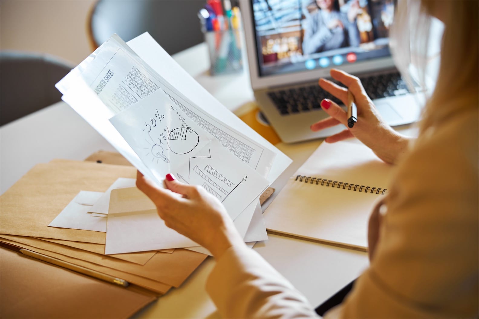 A person holding a document with charts and graphs is seated at a desk, immersed in a video call on their laptop. Envelopes, papers, and a notebook lie nearby. The focus on the hand with the pen suggests virtual CFO services Australia are being discussed for enhancing business strategies.