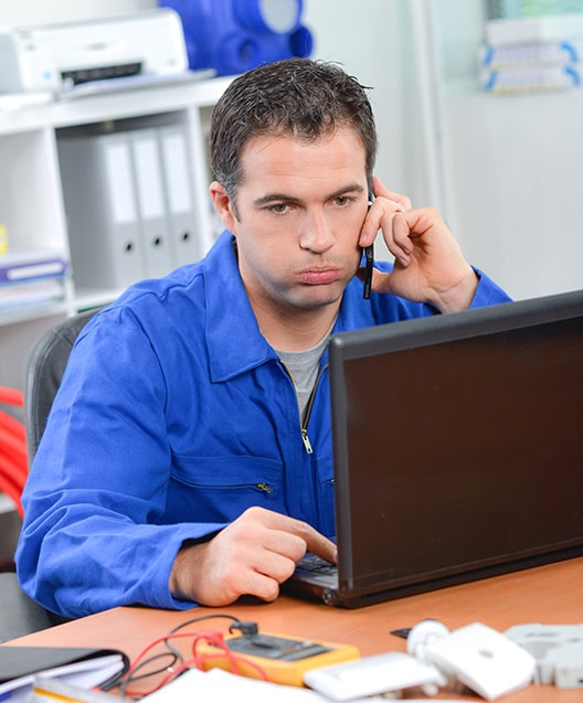 A person in a blue work uniform sits at a desk using a laptop, holding a phone to their ear, appearing focused and slightly stressed. Surrounded by tools and papers, they manage professional bookkeeping services. Shelves with files and other office items are visible in the background.