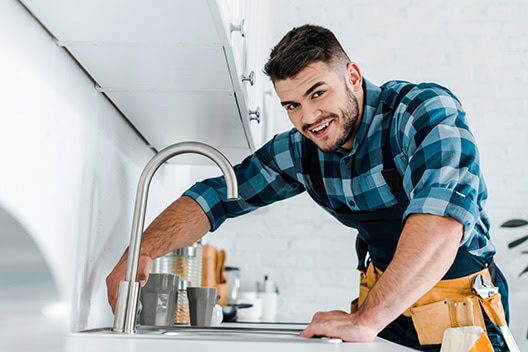 A man in a blue plaid shirt is fixing a kitchen faucet with precision, leaning over a white countertop. Focused, he uses a wrench from his tool belt in the sleek, modern kitchen—a scene as meticulous as professional bookkeeping services.