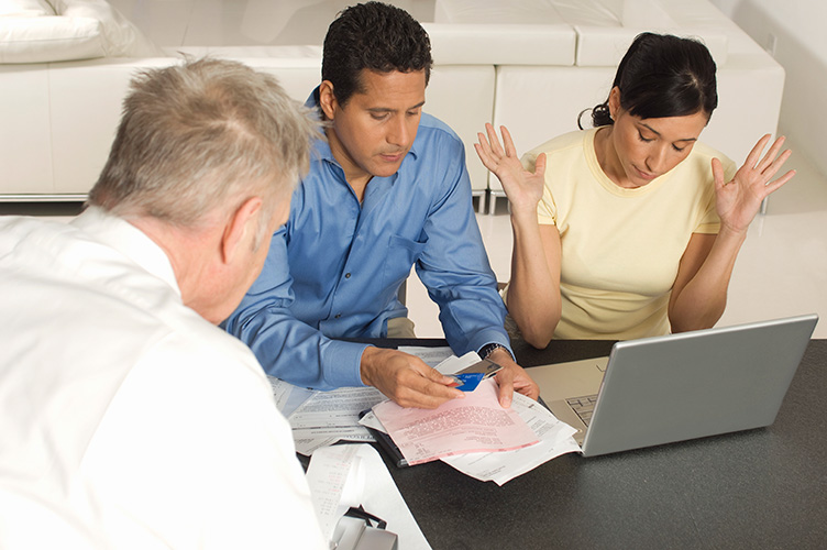A man and woman sit at a table with papers and a laptop, looking stressed; the woman raises her hands in frustration while another man in a white shirt sits across from them, offering help with ATO debts as they review documents.