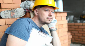 A construction worker wearing a yellow hard hat and gloves sits against a stack of bricks, holding a clipboard and looking thoughtful—perhaps considering the best business structure for tradies for his next project.
